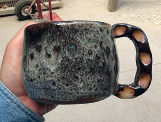 Speckled dark-glazed mug with a pierced knuckle-style handle, held in hand.