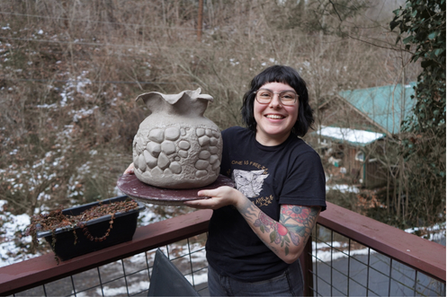 Breez Clifford on a mountain deck, holding a large raw coil-built pot.
