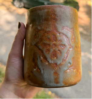 Tumbler carved with a medallion motif under a rust-and-sage iron glaze, held in hand outdoors.